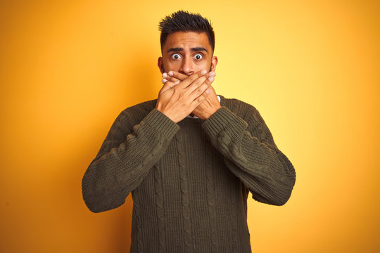 Young indian man wearing green sweater and shirt standing over isolated yellow background shocked covering mouth with hands for mistake. Secret concept.
