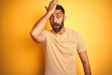 Young indian man wearing t-shirt standing over isolated yellow background surprised with hand on...