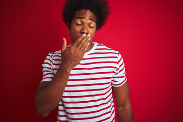 Young african american man with afro hair wearing striped t-shirt over isolated red background bored yawning tired covering mouth with hand. Restless and sleepiness.