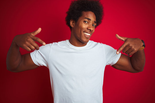 Young american man with afro hair wearing white t-shirt standing over isolated red background looking confident with smile on face, pointing oneself with fingers proud and happy.