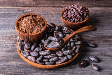 Whole dry cocoa beans, powder, nimbs in wooden bowls on old rustic background.
