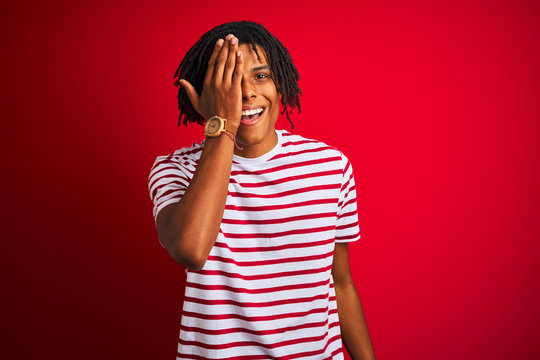 Young afro man with dreadlocks wearing striped t-shirt standing over isolated red background covering one eye with hand, confident smile on face and surprise emotion.