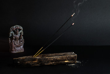 A burning, smoldering, incense stick with smoke, in a wooden stand, with a statuette of the Indian god Krishna, on a dark background.