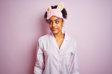 Young african american woman wearing pajama and mask over isolated pink background smiling looking to the side and staring away thinking.