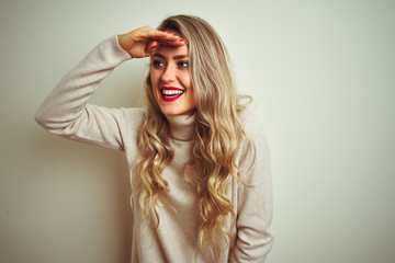 Beautiful woman wearing winter turtleneck sweater over isolated white background very happy and smiling looking far away with hand over head. Searching concept.