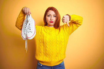 Young beautiful redhead woman holding white sneakers over yellow isolated background with angry...