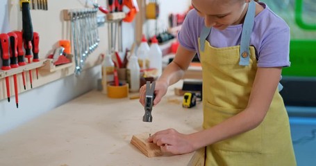 Girl 10 years old hammer a nail into a piece of wood in the wood workshop