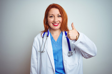 Young redhead doctor woman using stethoscope over white isolated background doing happy thumbs up gesture with hand. Approving expression looking at the camera showing success.