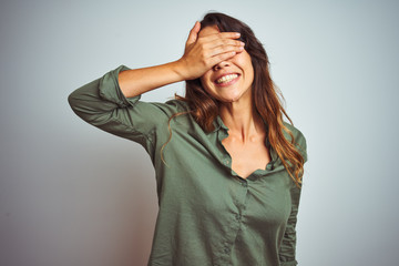 Young beautiful woman wearing green shirt standing over grey isolated background smiling and laughing with hand on face covering eyes for surprise. Blind concept.