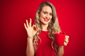 Young beautiful woman drinking a cup of cooffe over red isolated background doing ok sign with fingers, excellent symbol