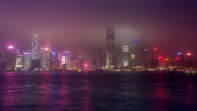 Timelapse Hong Kong boats sail against Central and Western district illuminated buildings and skyscrapers in dense mist at night zoom out