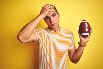 Young rugby player man holding a football ball over yellow isolated background stressed with hand on head, shocked with shame and surprise face, angry and frustrated. Fear and upset for mistake.