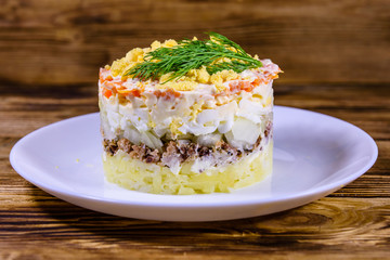 Plate with traditional russian salad Mimosa on a wooden table. Layered salad with potatoes, sardine, cheese, carrot, eggs and mayonnaise