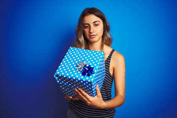 Young woman holding birthday present over blue isolated background stressed with hand on head, shocked with shame and surprise face, angry and frustrated. Fear and upset for mistake.