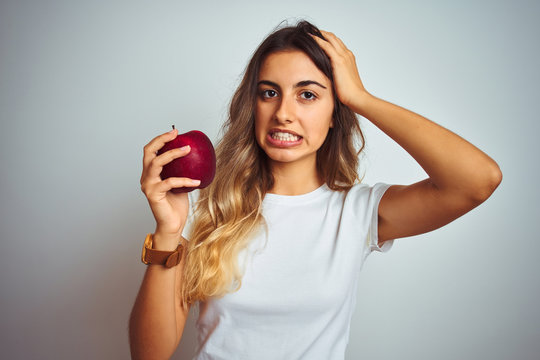 Young beautiful woman eating red apple over grey isolated background stressed with hand on head, shocked with shame and surprise face, angry and frustrated. Fear and upset for mistake.