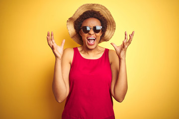 African american woman wearing summer hat and sunglasses over yellow isolated background celebrating mad and crazy for success with arms raised and closed eyes screaming excited. Winner concept