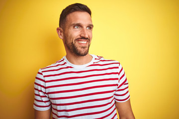 Fototapeta premium Young handsome man wearing casual red striped t-shirt over yellow isolated background looking away to side with smile on face, natural expression. Laughing confident.