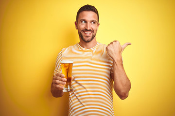 Young handsome man drinking a pint glass of beer over isolated yellow background pointing and showing with thumb up to the side with happy face smiling