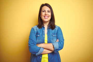 Young beautiful brunette woman wearing casual denim jacket over yellow isolated background happy face smiling with crossed arms looking at the camera. Positive person.