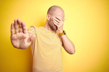 Young bald man with beard wearing casual striped t-shirt over yellow isolated background covering...