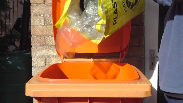 Slow motion close POV shot of a man opening a bin lid then pouring various household plastic items from a yellow portable recycle carrier into an orange recycle wheelie bin, then closing the lid.