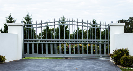 Grey metal wrought iron driveway property entrance gates set in white concrete brick fence, garden...