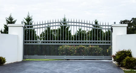 Fotobehang Grey metal wrought iron driveway property entrance gates set in white concrete brick fence, garden trees in background © squirrel7707