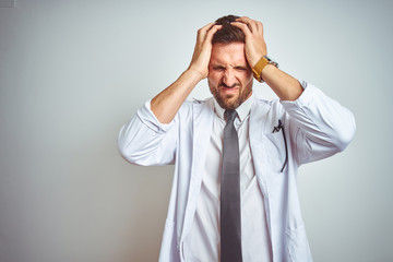 Young handsome doctor man wearing white profressional coat over isolated background suffering from headache desperate and stressed because pain and migraine. Hands on head.
