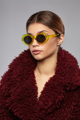 Close-up shot of a young brunette European lady in burgundy-coloured furs and oversized sunglasses with black oval lens and wide transparent yellow rim. The photo is made on the gray background.