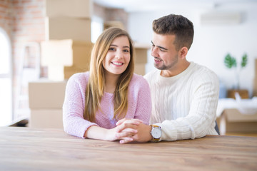 Young beautiful couple sitting on the table at home, hugging in love very happy for moving to new home with cardboard boxes behind them