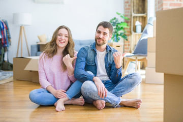Fototapeta na wymiar Young beautiful couple moving to a new house sitting on the floor smiling with happy face looking and pointing to the side with thumb up.