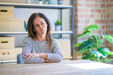 Middle age senior woman sitting at the table at home puffing cheeks with funny face. Mouth inflated with air, crazy expression.