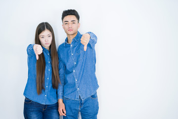 Beautiful young asian couple over white isolated background looking unhappy and angry showing rejection and negative with thumbs down gesture. Bad expression.