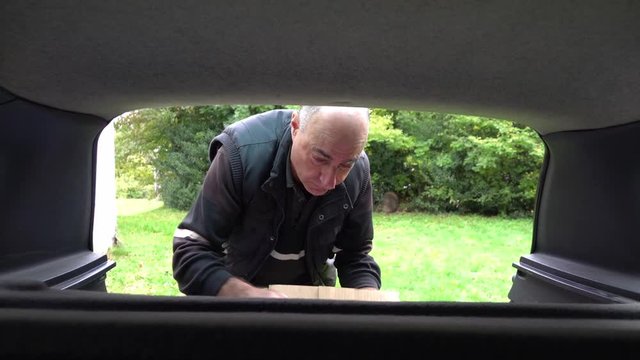 Serious mature bald man folds a cardboard box in the trunk of a car and picks it up