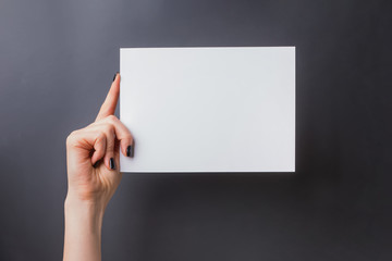 Woman's hand with black nailpolish holding a blank paper against black wall