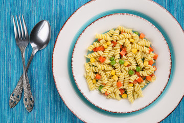 Top down, flat lay of delicious, vegetarian fusilli pasta with basil pesto, sweet corn, carrot and peas in a plate on a turquoise blue tablecloth with spoon and fork