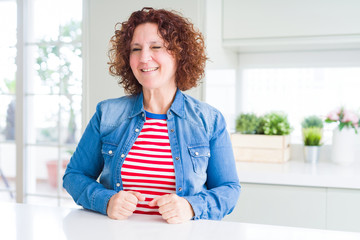 Middle age senior woman with curly hair wearing denim jacket at home winking looking at the camera...