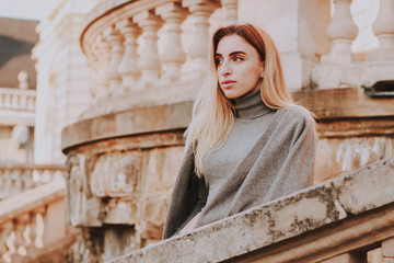 Stylish autumn fashionable girl image. Gray blazer, pleated skirt and gray coat. Blond woman/Old city background.