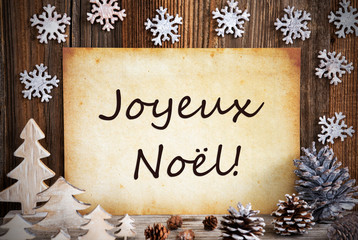 Obraz na płótnie Canvas Old Paper With French Text Joyeux Noel Means Merry Christmas. Christmas Decoration Like Tree, Fir Cone And Snowflakes. Brown Wooden Background