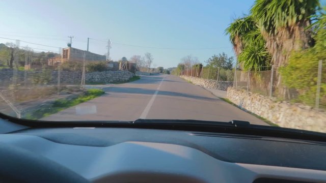 Drive on a winding road through the village with a net fencing on a short wall and surrounded with trees and green field, electric polls and a telecom tower, Captured from windscreen of the car.
