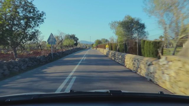 Drive on a winding road with a wired fencing on a short wall and surrounded with trees and green field under the blue sky.