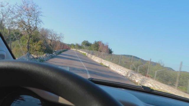 A close up view of a steering and dashboard of a car with drive on a winding road through the village with a net fencing on a short wall and surrounded with trees and green field.