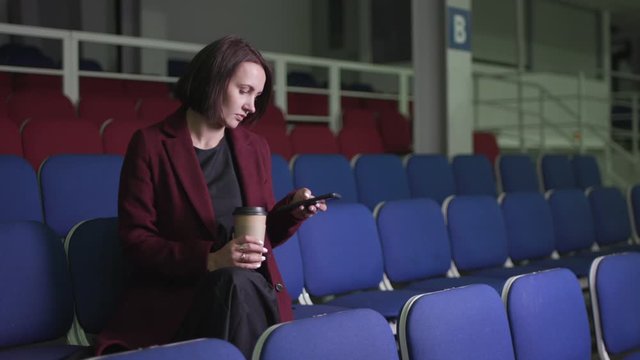 Woman sitting alone in empty stadium checking smartphone and holding coffee cup