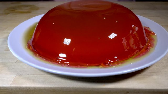 Slow motion close POV dolly shot of a whole wobbling strawberry jelly / jello being turned out of a glass bowl, to rest on a plate on a kitchen worktop, ready to be served.