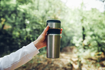 Unrecognizable woman traveler holding thermos mug on background of green forest. Travel, hiking,...