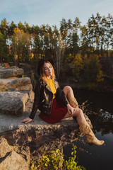 Woman in a leather jacket and hat on the edge of a cliff near the lake in the autumn forest