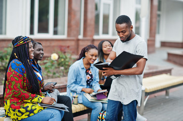 Group of five african college students spending time together on campus at university yard. Black afro friends studying at bench with school items, laptops notebooks.