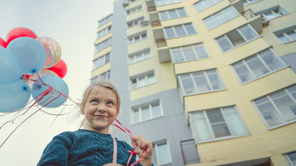 A charming little girl in a sweater smiles and holds colorful balloons on the background of a residential multi-storey building.