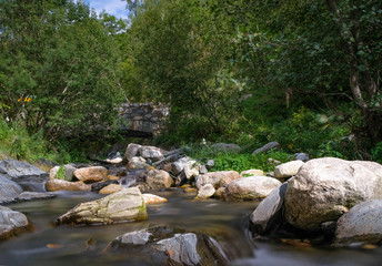 Naklejka na ściany i meble Ancient bridge over a mountain stream. Mountain river in the forest on blue sky and green trees background. Forest landscape with water movement between large stones. Long exposure.