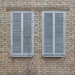 Two windows with closed grey metal shutters on dirty brick wall background